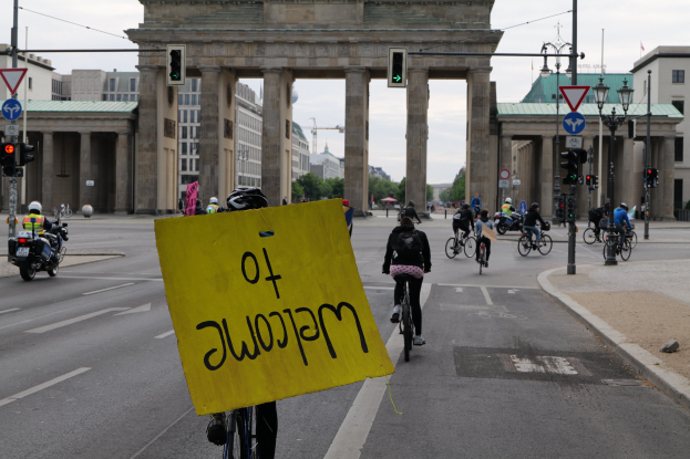 Eine Gruppe von Menschen, die Fahrräder am Brandenburger Tor in Berlin vorbeifährt, einer hält ein gelbes Schild, mit Laternenmasten, Verkehrsampeln, Gebäuden, Bäumen und einem klaren Himmel im Hintergrund.