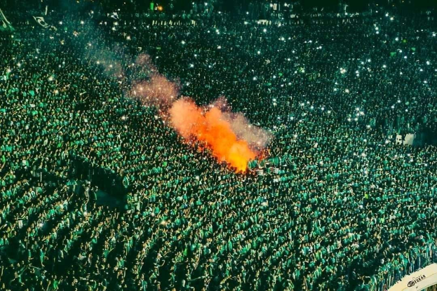 Luftaufnahme einer großen Menge in einem Stadion mit einem zentralen Feuer, das die Umgebung erhellt.