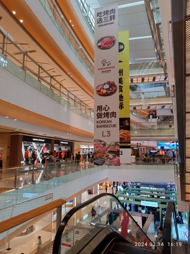 Ein Blick ins Innere eines Einkaufszentrums mit einer Rolltreppe in der Mitte, Menschen mit Handtaschen, von der Decke hängende Banner und Geschäfte im Hintergrund.