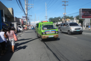 Ein grüner Lieferwagen fährt auf einer Straße mit hohen Gebäuden auf beiden Seiten, Fußgängern auf dem Gehweg, Strommasten mit Drähten, Bäumen im Hintergrund und einem klaren blauen Himmel.