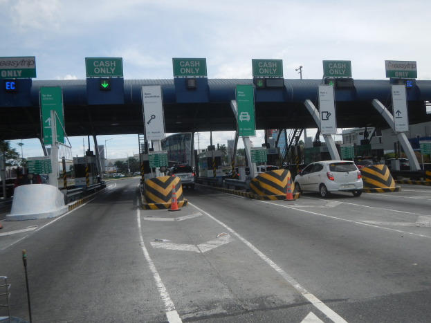 Mautstation mit darunterfahrenden Autos, umgeben von Verkehrskegeln, Absperrpollern, Hinweisschildern, einem Bogen, Laternenmasten, Bäumen, Gebäuden und einem bewölkten Himmel.
