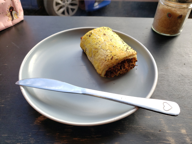 Ein Teller mit einem Stück Essen und einem Messer daneben, ein Glasgefäß auf dem Tisch und ein Fahrzeug durch die Glasscheibe im Hintergrund.