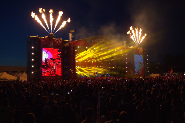 Eine große Menschenmenge auf einem Musikfestival schaut auf einer hell erleuchteten Bühne Performern zu, mit Zelten links und Feuerwerk am dunklen Himmel dahinter.