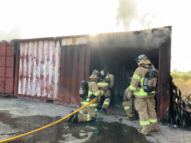 Feuerwehrleute in Helmen und Schutzausrüstung löschen ein Feuer in einem raucherfüllten Seecontainer, mit Bäumen und Himmel im Hintergrund.
