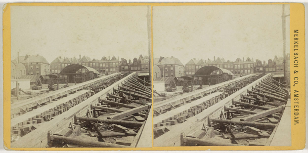 Schwarz-weiß-Foto einer Bahnschiene mit Gebäuden im Hintergrund, Text 'Merkelbach & Co, Co. Amsterdam' rechts und verstreute hölzerne Gegenstände im Vordergrund.