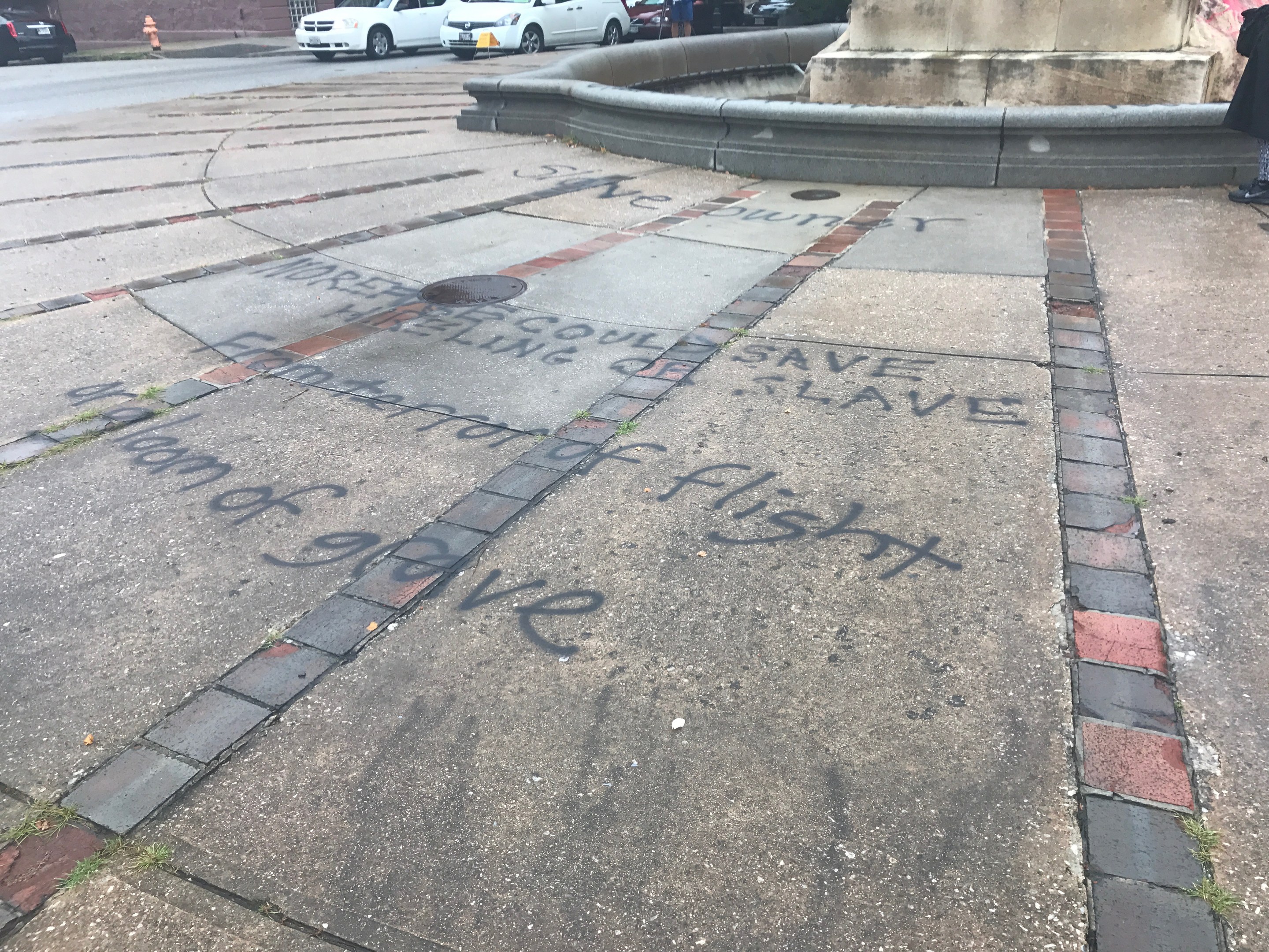 Schwarze Graffiti-Schriftzug "Black Lives Matter" auf einem Gehweg vor einem Gebäude, mit Fahrzeugen, Menschen, Bäumen, Masten und einer Wand in der Nähe.
