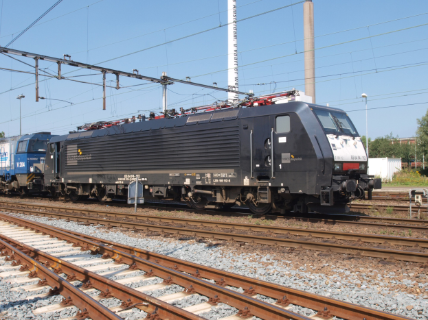 Schwarzer Zug fährt auf Schienen neben einem Wald mit Strommasten und Drähten, Bäumen, Gebäuden und einem klaren blauen Himmel im Hintergrund.