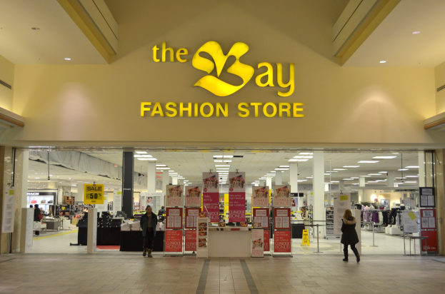 Außenansicht eines Bay Fashion Stores in einem Einkaufszentrum mit Menschen, die gehen, Texttafeln an Wänden, Säulen, Deckenleuchten und Hintergrundanzeigen.