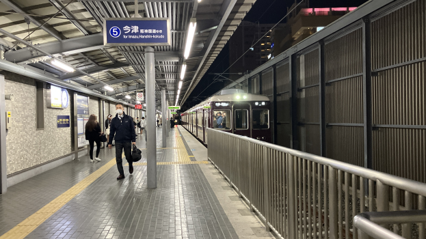 Ein Zug fährt auf ein Bahnsteiggleis zu, mit Menschen in der Nähe, einem Geländer auf der rechten Seite, Gebäuden im Hintergrund, Bahnsteigbeleuchtung und einer Anzeigetafel.