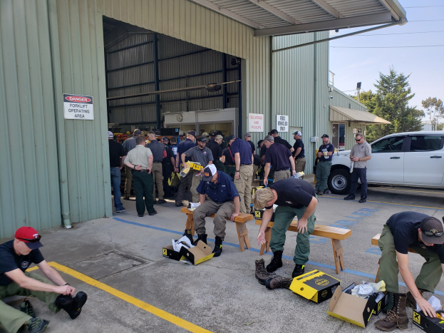 Eine Gruppe von Menschen bei einer Ausbildungssitzung der Nationalgarde, einige sitzen auf Bänken und andere stehen, mit verstreuten Kartonboxen, Schuhen und Gegenständen vor einem Gebäude, vor einer Kulisse aus Fahrzeugen, Bäumen, Gebäuden, Polen, Drähten und einem klaren blauen Himmel.