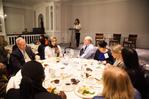 Eine Gruppe von Menschen sitzt um einen Esstisch mit Tellern, Bestecken und Trinkgefäßen, während eine Frau in der Nähe eines Podiums mit einem Mikrofon im Hintergrund an einem Event mit Stühlen, Tischen, Flaschen und Wandlampen steht.