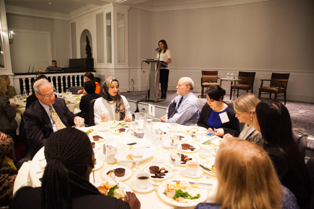 Eine Gruppe von Menschen sitzt um einen Esstisch mit Tellern, Bestecken und Trinkgefäßen, während eine Frau in der Nähe eines Podiums mit einem Mikrofon im Hintergrund an einem Event mit Stühlen, Tischen, Flaschen und Wandlampen steht.