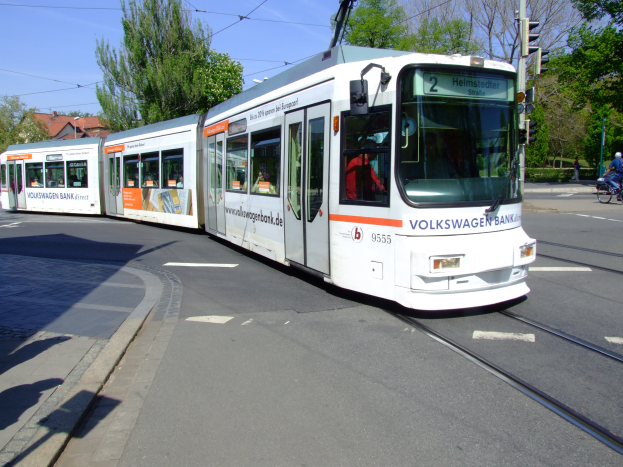 Eine weiße und orangefarbene Straßenbahn befindet sich auf einer Stadtstraße mit einem Fahrradfahrer auf der rechten Seite, Bäumen, Gebäuden und einem klaren blauen Himmel im Hintergrund.