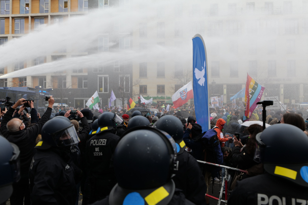 Polizeibeamte in Einsatzausrüstung stehen vor einer Menge mit Kameras und Helmen, während ein Wasserwerfer die Menge bespritzt; Gebäude, Bäume und ein blauer Himmel sind im Hintergrund sichtbar.