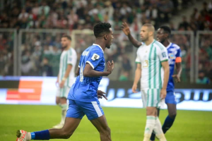 Ein Fußballspieler in einem blauen und weißen Trikot feiert nach einem Torerfolg, umgeben von einer Gruppe von Menschen in weißen und grünen Trikots, mit einer weißen und blauen Tafel und einem Geländer im unscharfen Hintergrund.