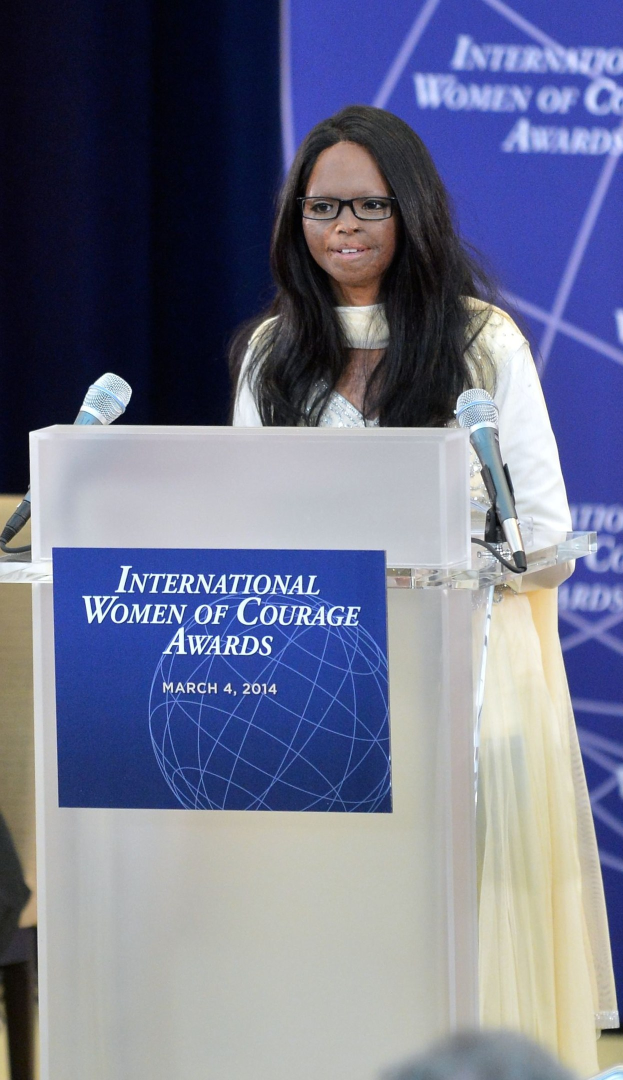 Frau In Weißen Kleid Und Brille, Die Auf Einem Podium Mit Zwei Mikrofonen, Einer Tafel Und Einem Blauen Banner Mit Der Aufschrift "Internationale Frauen Des Mutes Awards 2014" Hinter Ihr Spricht.