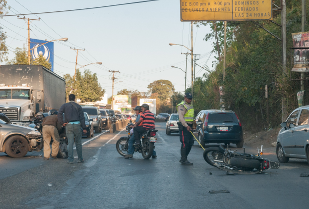 Gruppe von Menschen um ein verunglücktes Motorrad auf der Seite einer Straße mit mehreren Fahrzeugen, einschließlich eines Lastwagens, und Hintergrundelementen wie Bäume, Pfosten, Lampen, Schilder und Himmel.