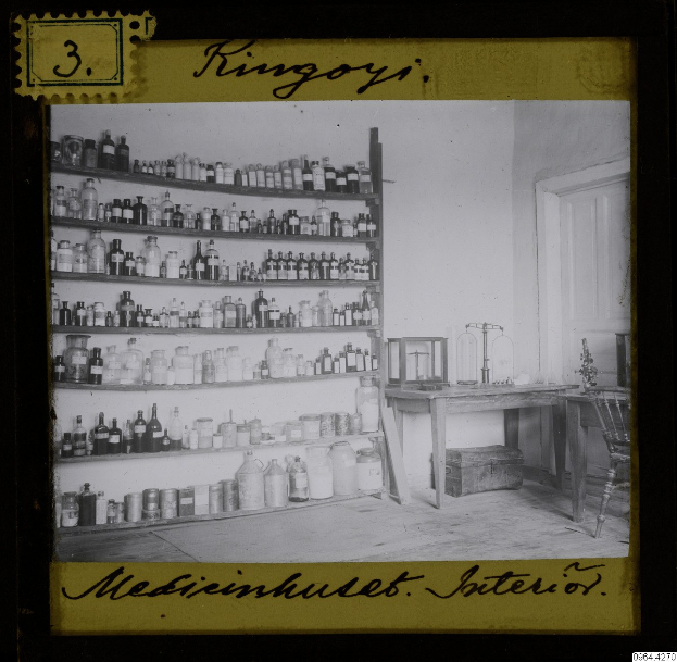 Schwarz-weiß-Foto eines vintage-Apothekeninneren mit Regalen voller Flaschen, einem Tisch mit Gegenständen, einem Stuhl, einer Tür und einer Wand mit Text am unteren Rand.