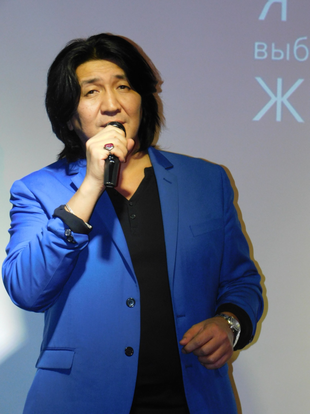 Ein Mann in einem blauen Anzug mit einem ernsten Gesichtsausdruck hält ein Mikrofon und steht vor einem Bildschirm mit Text, gekleidet in einen schwarzen Anzug und ein weißes Hemd.