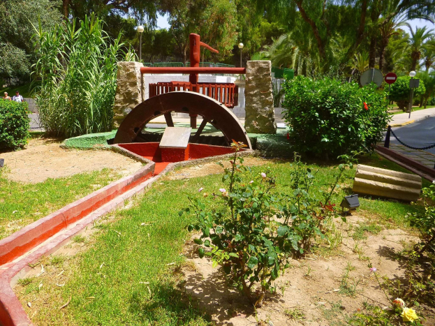 Minigolfanlage in einem Park mit Grünfläche, einer Straße mit Fußgängern, Schilderpfosten und einem klaren Himmel im Hintergrund.