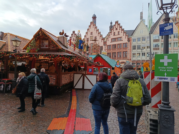 Menschen spazieren auf einem Kopfsteinpflasterweg neben einem Weihnachtsmarkt in Nürnberg, Deutschland, mit Laternenmasten, Texttafeln und Gebäuden im Hintergrund unter einem bewölkten Himmel.