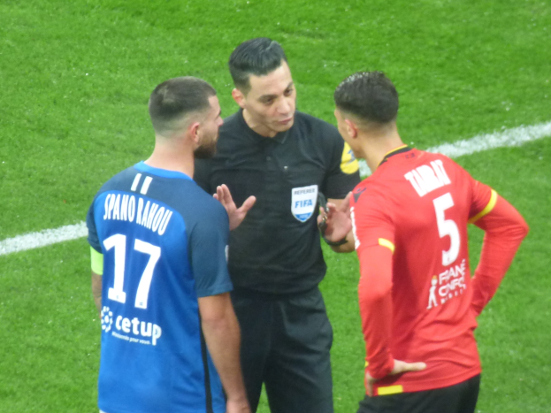 Schiedsrichter spricht mit zwei Männern auf einem Fußballfeld; einer trägt ein schwarzes Hemd und Hosen, der andere ein blaues und rotes T-Shirt mit Text und Brille.