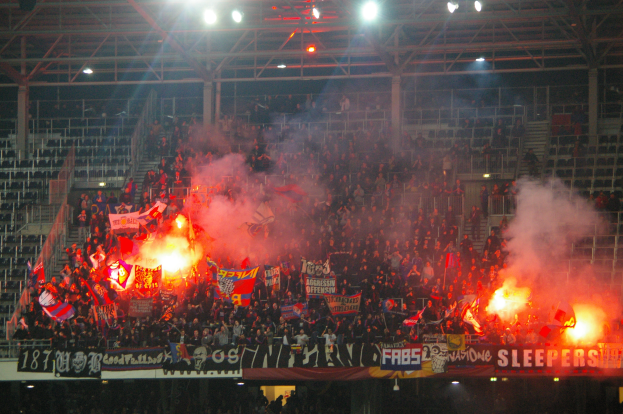 Eine große Menschenmenge in einem Stadion hält Fahnen und Banner, mit Leuchtraketen und Rauch, der eine lebendige Atmosphäre schafft, unter einer Decke mit Deckenleuchten und Metallrahmen.
