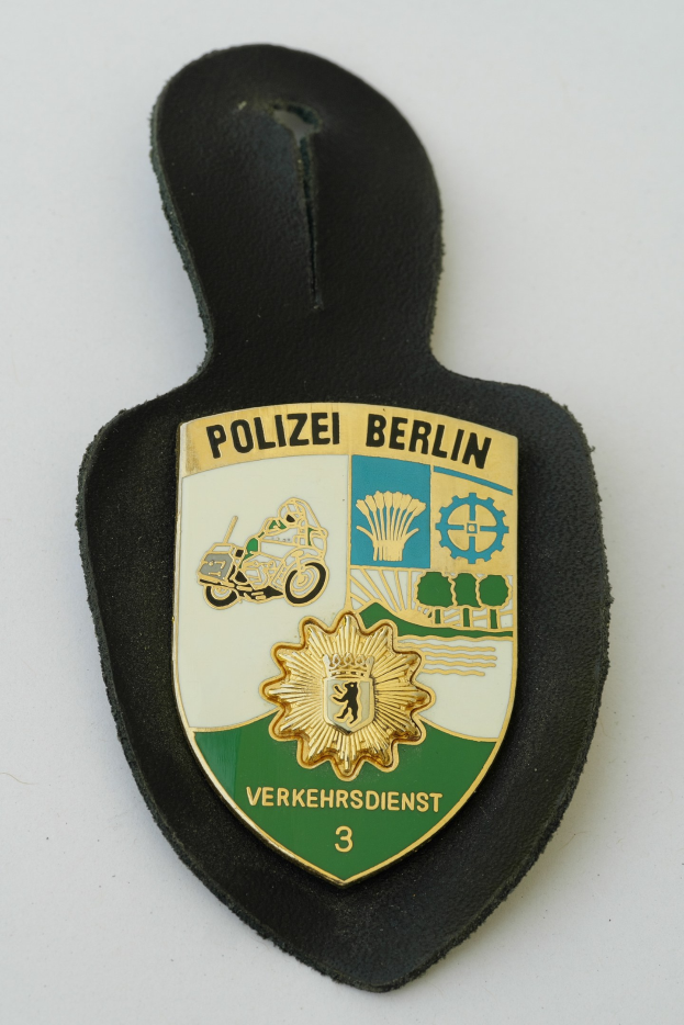 Ein schildförmiges Polizeibadge mit 'Police Berlin' in fetter schwarzer Schrift auf einem blauen Hintergrund mit einem weißen Stern und Rahmen, das auf einer Oberfläche liegt.