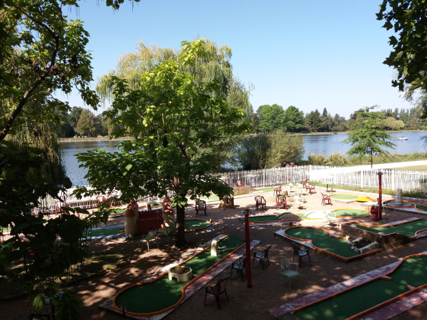Minigolfplatz in einem Park mit grünem Gras, Bäumen und einem Zaun, umgeben von verstreuten Stühlen und Tischen, einem Gewässer mit einem Boot im Hintergrund und sichtbarem Himmel.
