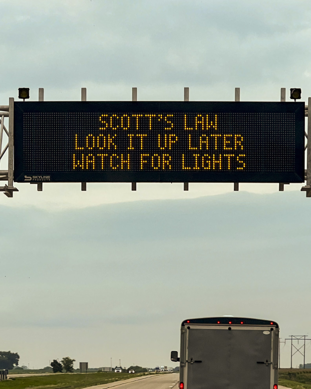 Ein Lkw fährt auf einer Autobahn mit einem Schild an der Seite, auf dem "Scott's Law: Look it Up Later, Watch for Lights" steht, umgeben von Gras, Bäumen und Pfählen, mit dem Himmel im Hintergrund.